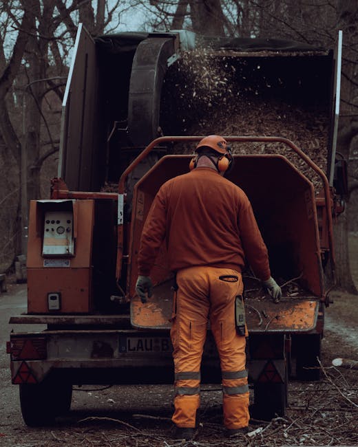 A gardener wearing an orange protective jacket, matching trousers with reflective stripes, gloves, and a helmet stands with their back to the camera, operating a large industrial wood chipper amidst a
